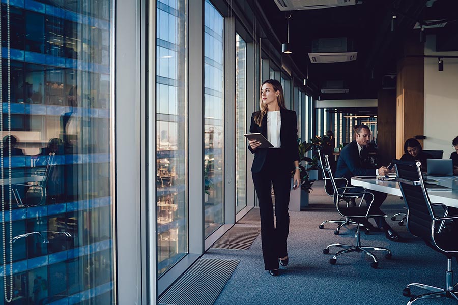 Business Insurance - Woman Looks Out the Window From a High Rise Office Building, Holding a Tablet, Employees at a Conference Table Behind Her