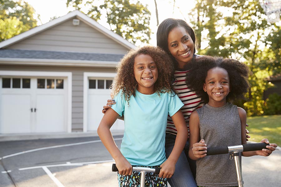 Personal Insurance - Mother and Young Daughters Smile in Their Driveway, Detached Double Garage Behind Them, the Girls Standing on Scooters